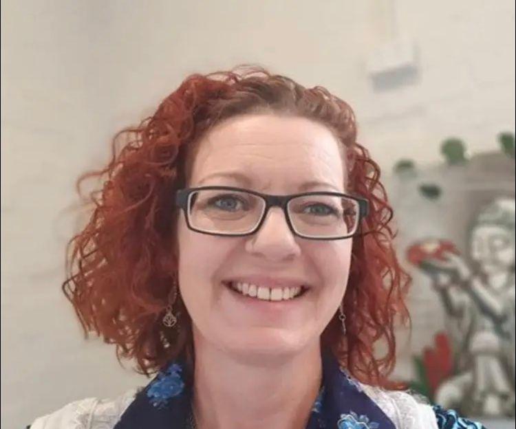 Lainey Bowler - a headshot of a lady with curly bobbed reddish auburn hair. Smiling warmly with a calm disposition, wearing glasses.