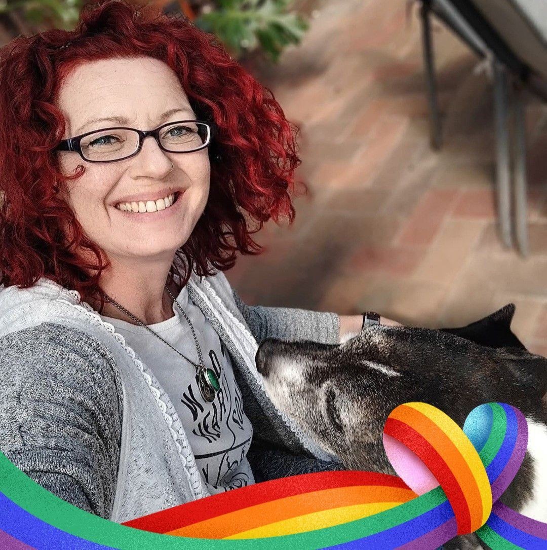 Smiling red haired lady, rainbow pride ribbons with a grey faced dog.